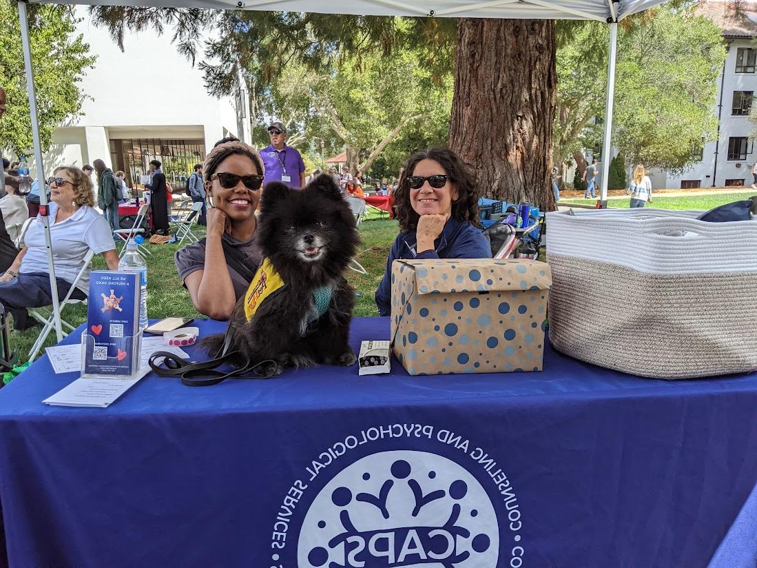 Wellness Fair picture with Joey the therapy dog and two CAPS staff members helping out at the table.