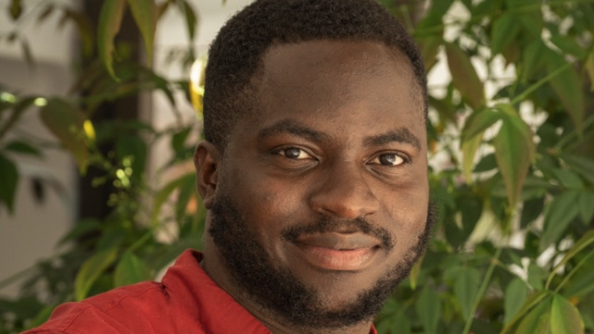 Kehinde Badiru smiles wearing a red shirt in front of trees